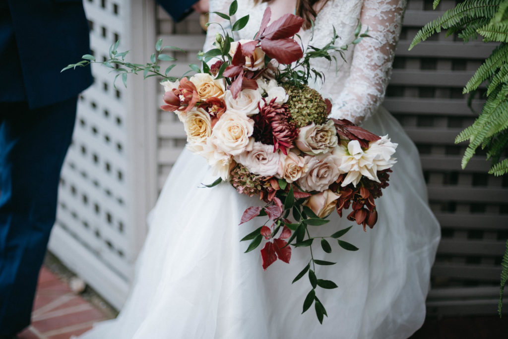 A bridal bouquet with roses, dahlias, hydrangea, and greenery in shades of mauve, white, burgundy, and orange.