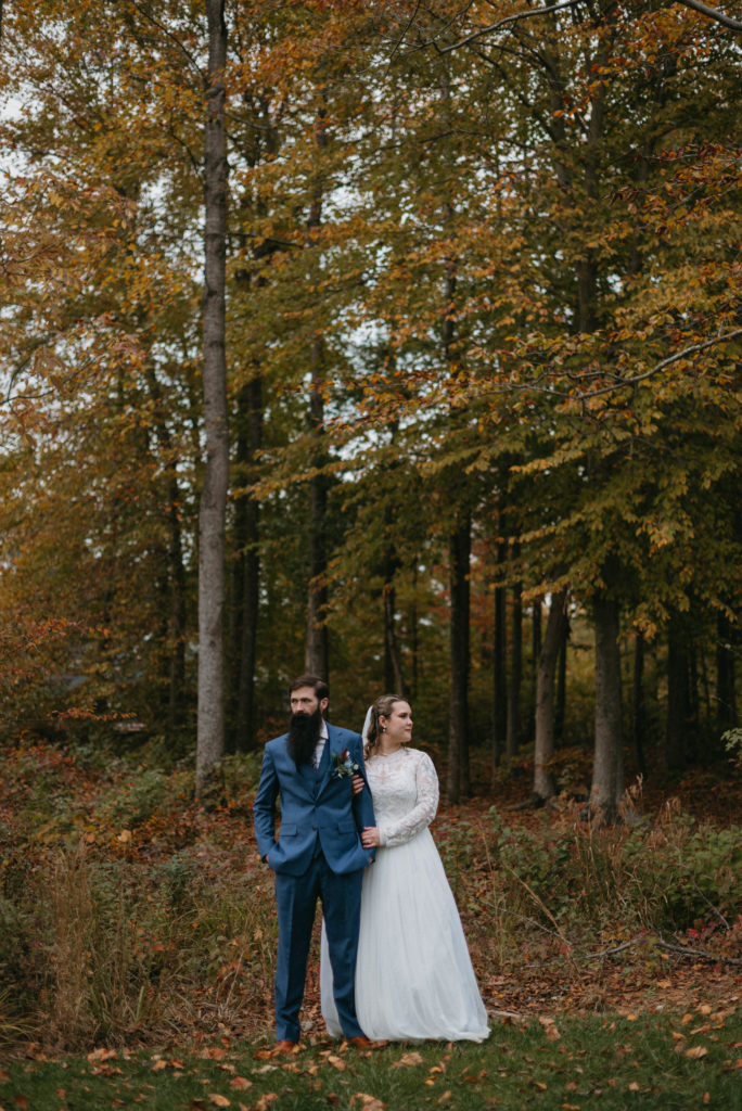 a couple in wedding attire stand beneath colorful trees and look in opposite directions. 