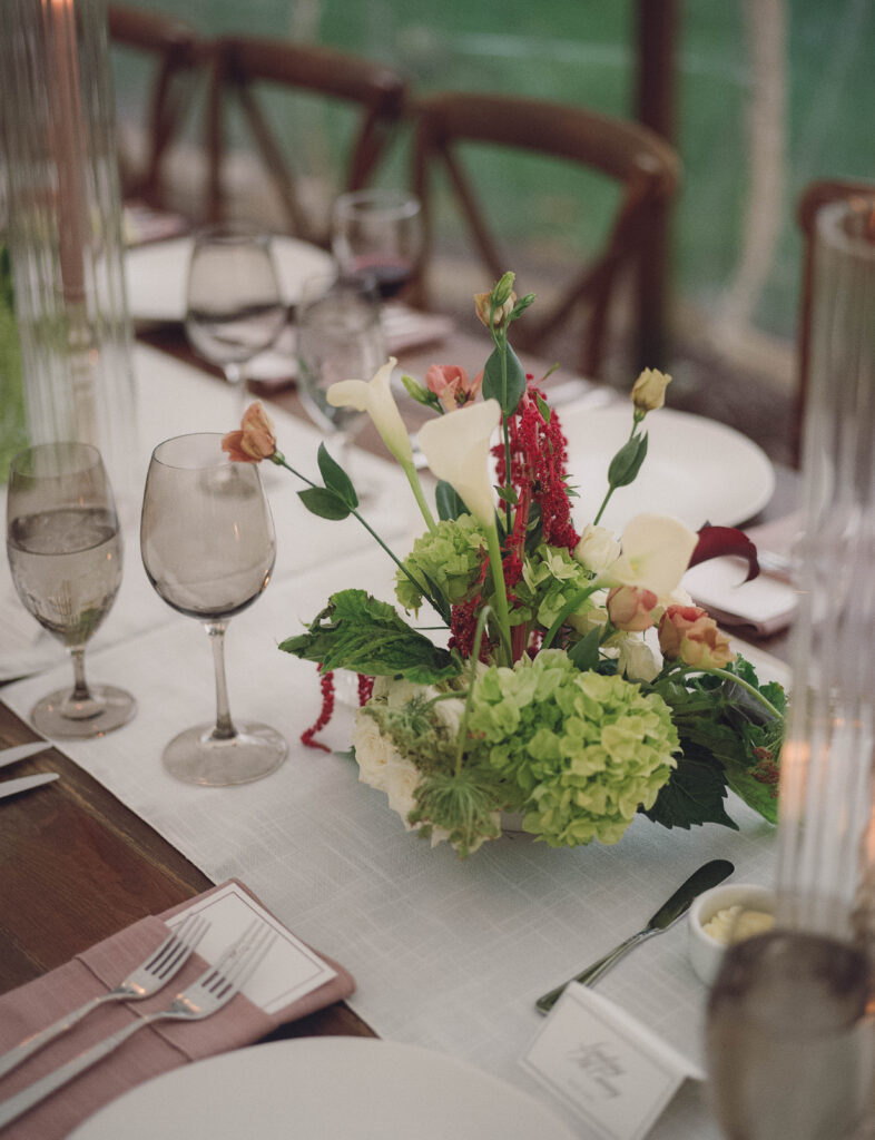 A green and maroon floral arrangement on a long table surrounded by candles and glassware