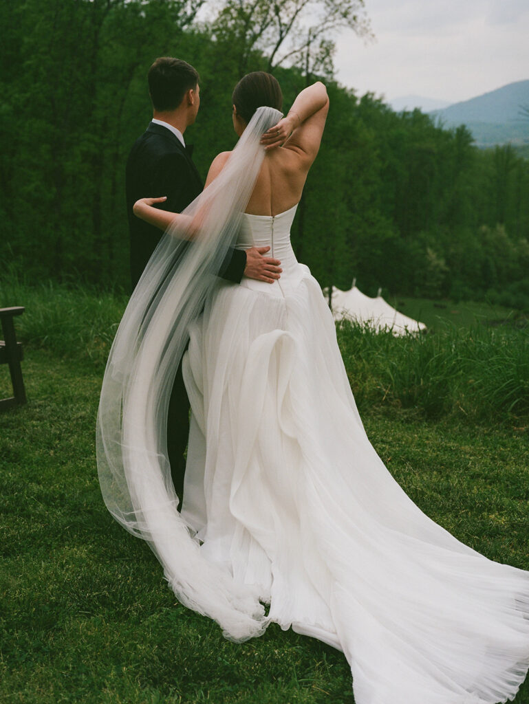 A bride and groom look over their wedding reception tent while up on a mountain. A gust of wind comes by surprise and the bride reaches back to secure her veil as it blows in the wind.