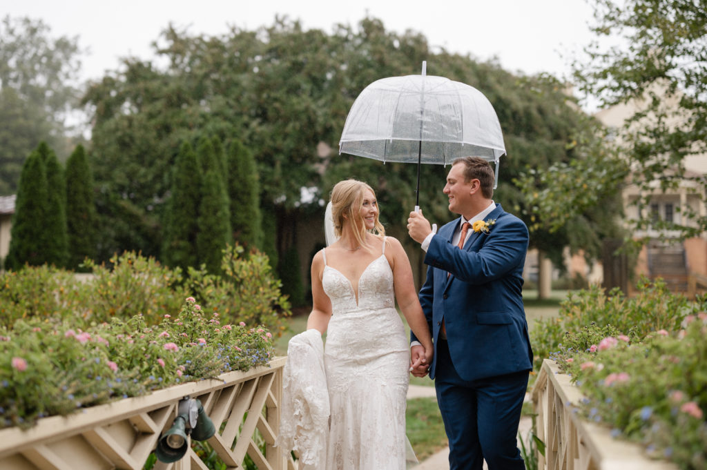 bride and groom hold hands and walk through a garden together in the rain with an umbrella