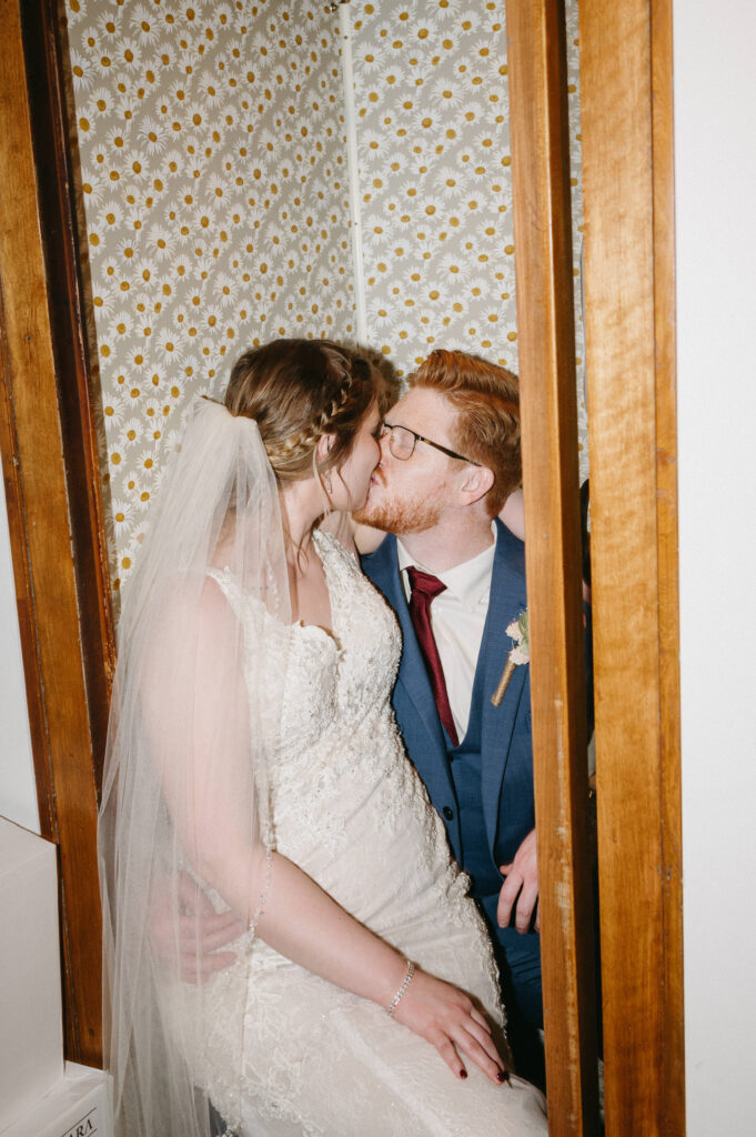 a couple kisses in an old phone booth covered in floral wallpaper