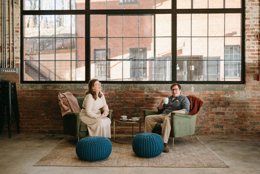 A couple sitting in chairs next to each other at a coffee shop with and exposed brick wall behind them. 