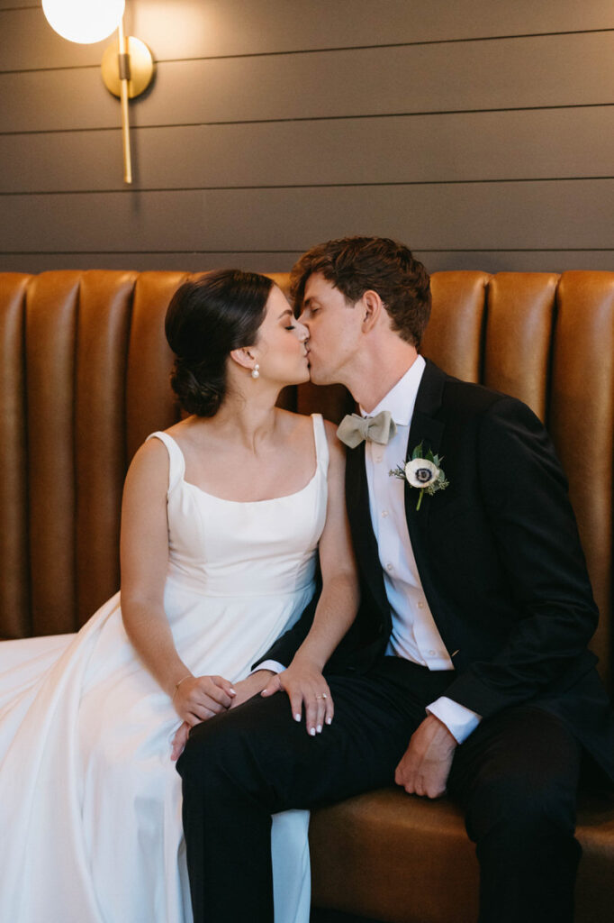 A wedding couple sitting on a tan leather couch kissing. 