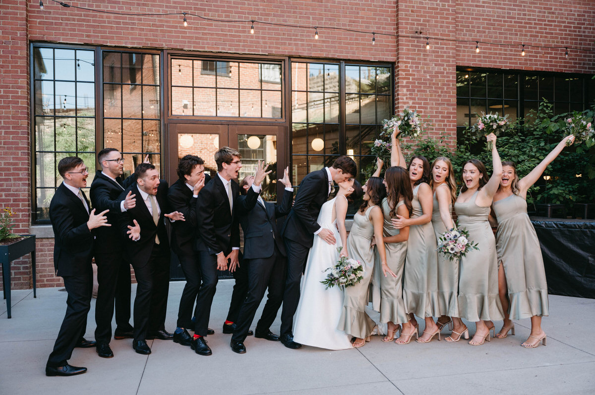 A bride and groom kissing with their wedding parties on either side of them at The Wool Factory wedding venue.