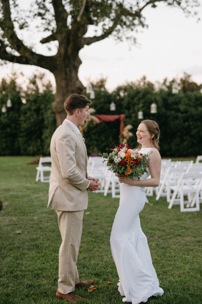 A bride and groom laughing as they talk to each other while standing on a lawn.