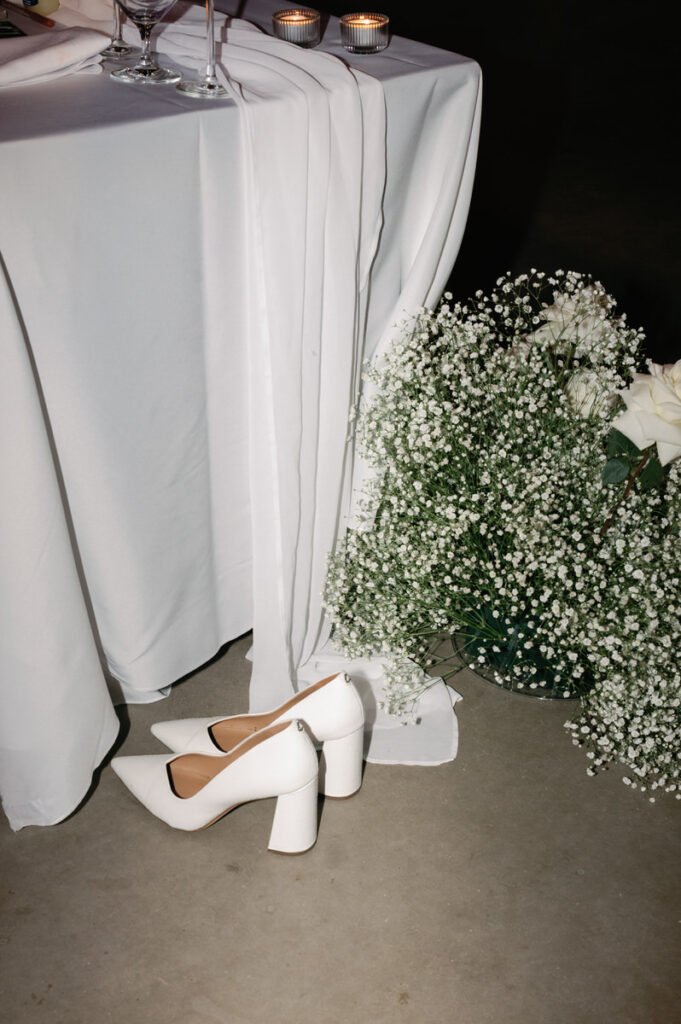 White heels sitting on the ground at the foot of a table next to a bouquet of flowers