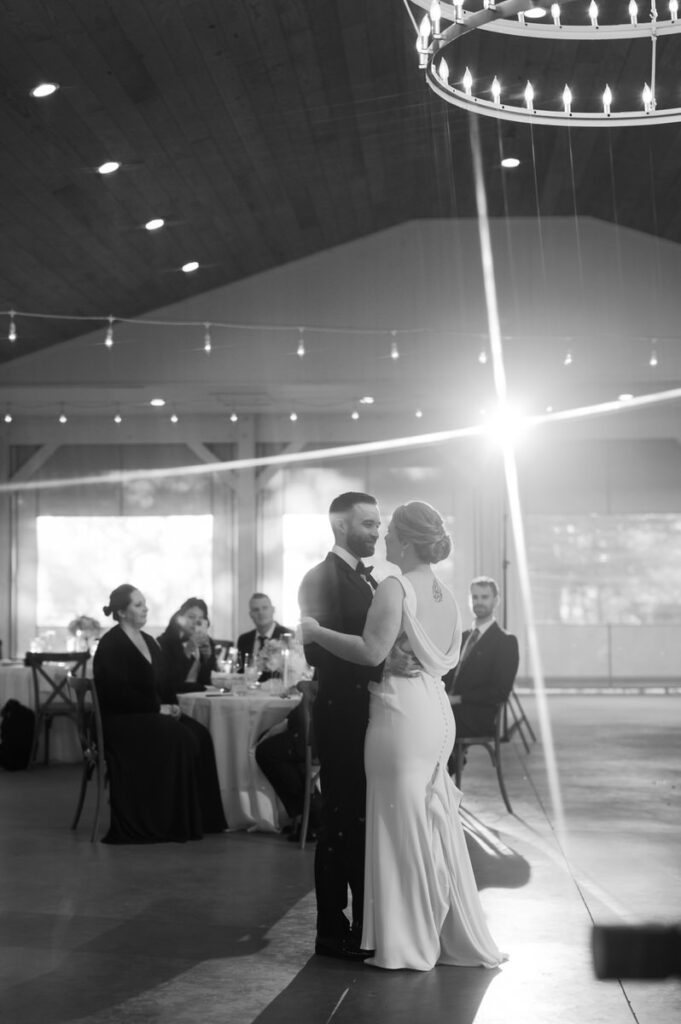 A black and white photo of a wedding couple during their first dance with their guests watching while sitting at tables. 