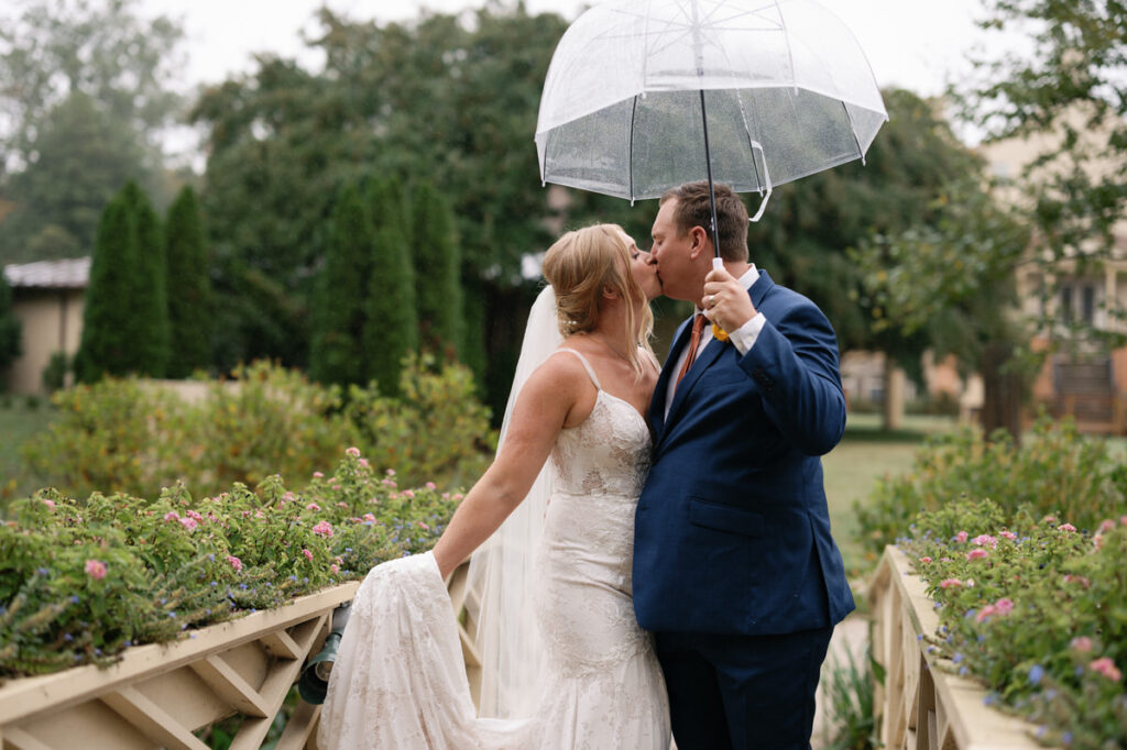 A bride and groom kissing while standing under a clear umbrella.