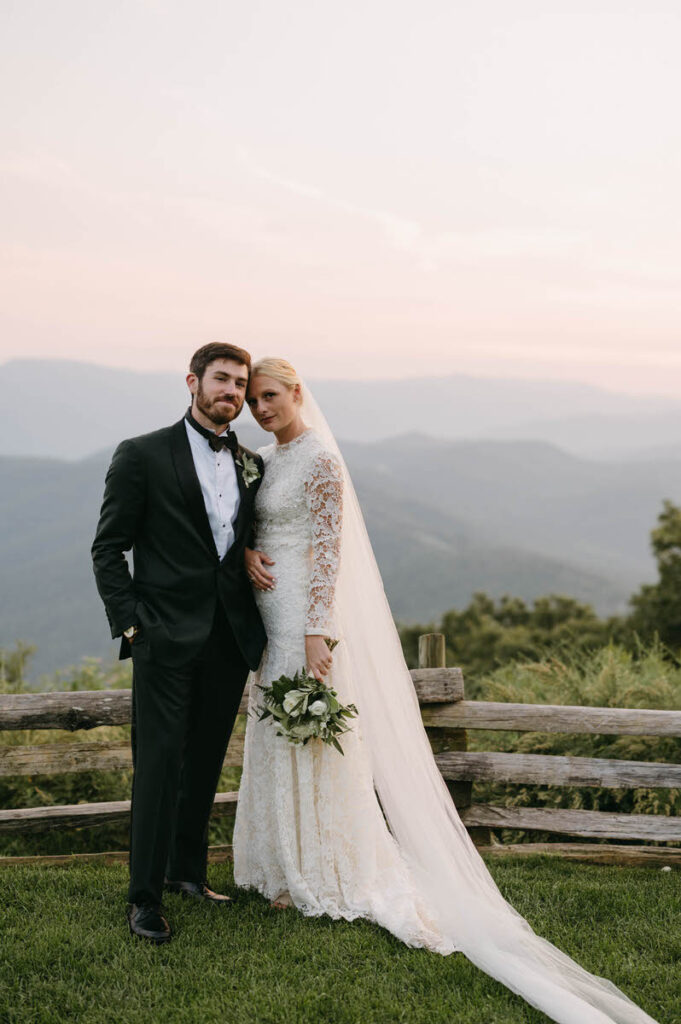 A wedding couple standing on the edge of a lookout smiling with a view of the mountains.  