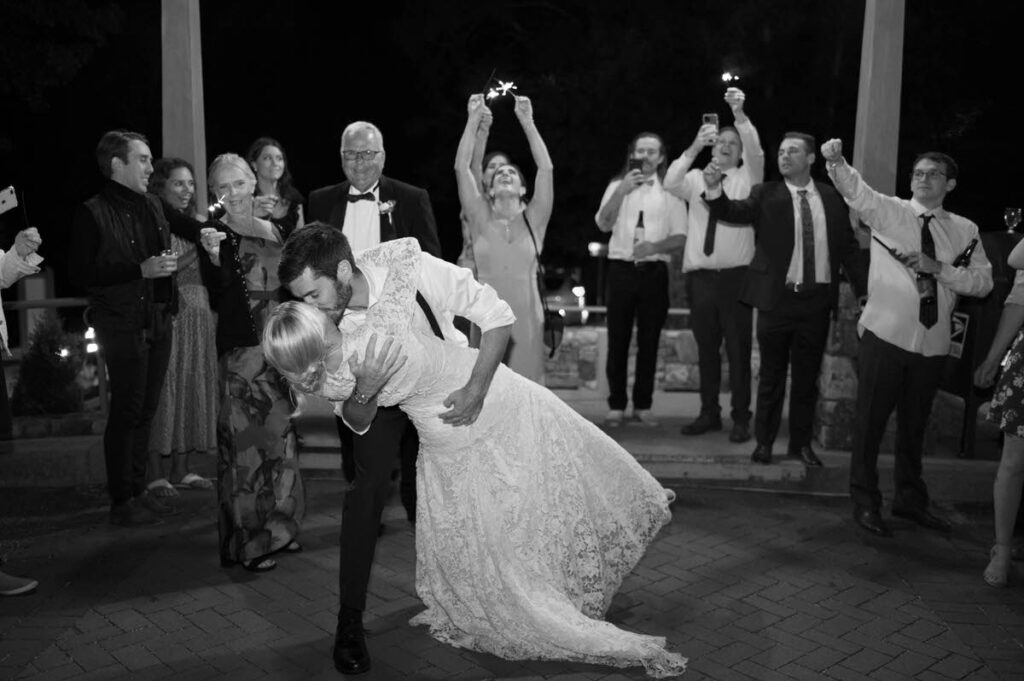 A groom dipping a bride as they kiss and wedding guests cheer behind them. 