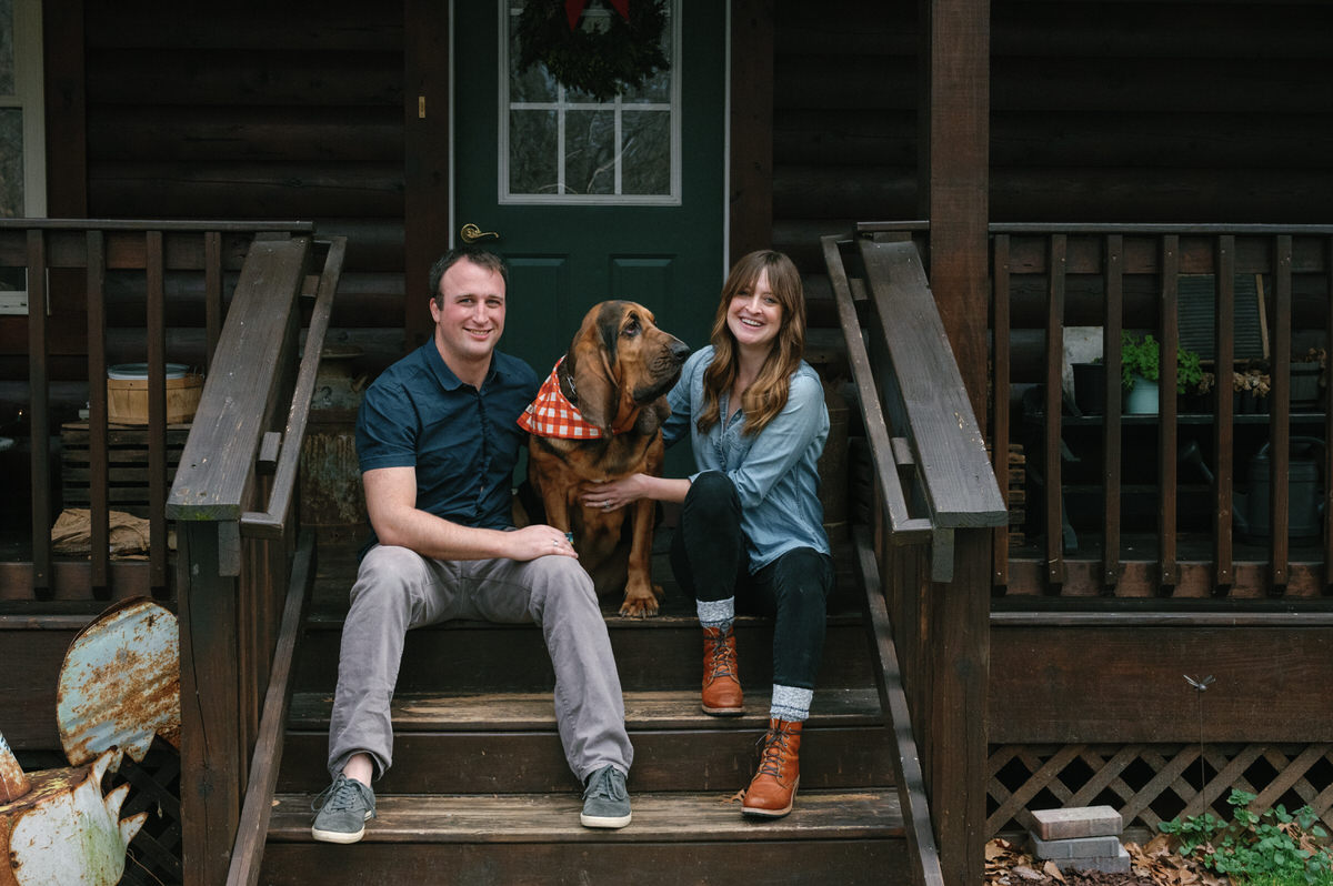 A couple sitting on a wooden staircase in front of an house with their dog showing off one style of casual engagement session outfit ideas.