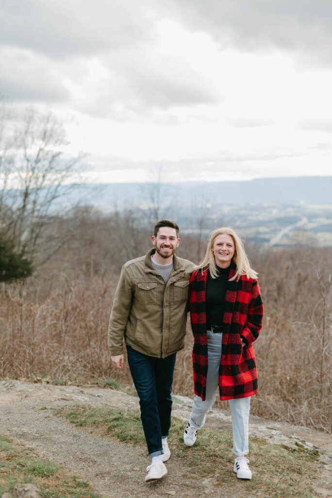 A couple smiling with their arms around each other as they walk in a wooded area in winter. 