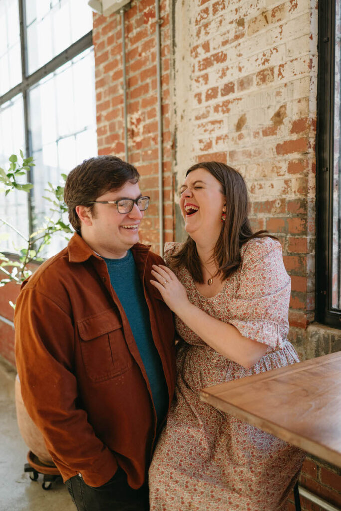 A woman sitting on a stool as she laughs and has her arm around her partner standing next to her. 