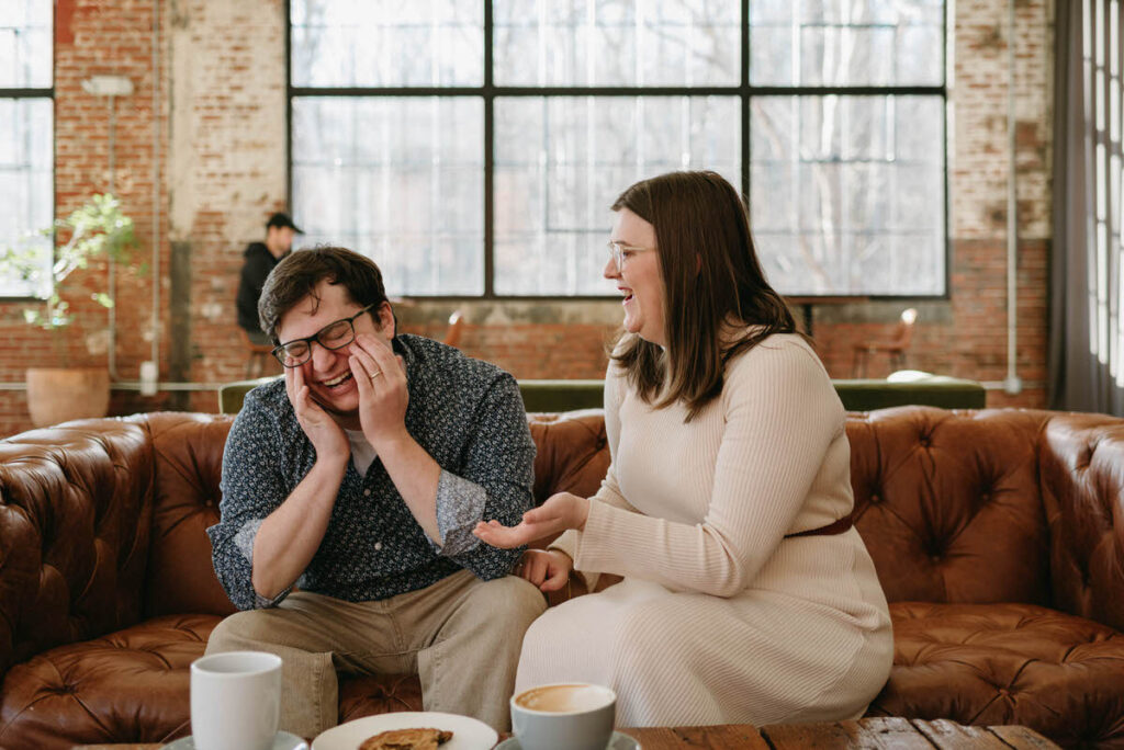 A couple laughing while sitting next to each other on a brown leather couch in a coffee shop. 