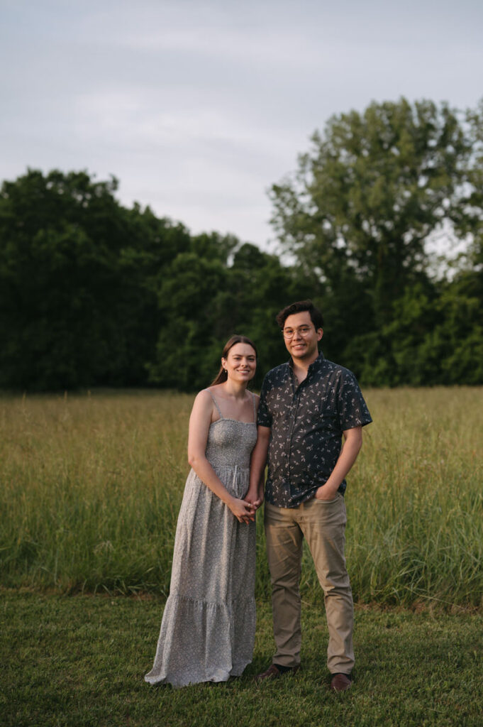 An engaged couple holding hands and smiling while standing in a field with overgrown grass and green trees behind them. 