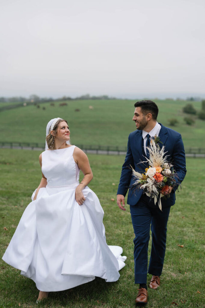 A wedding couple walking through a field while one holds their dress and the other holds a bouquet of flowers. 
