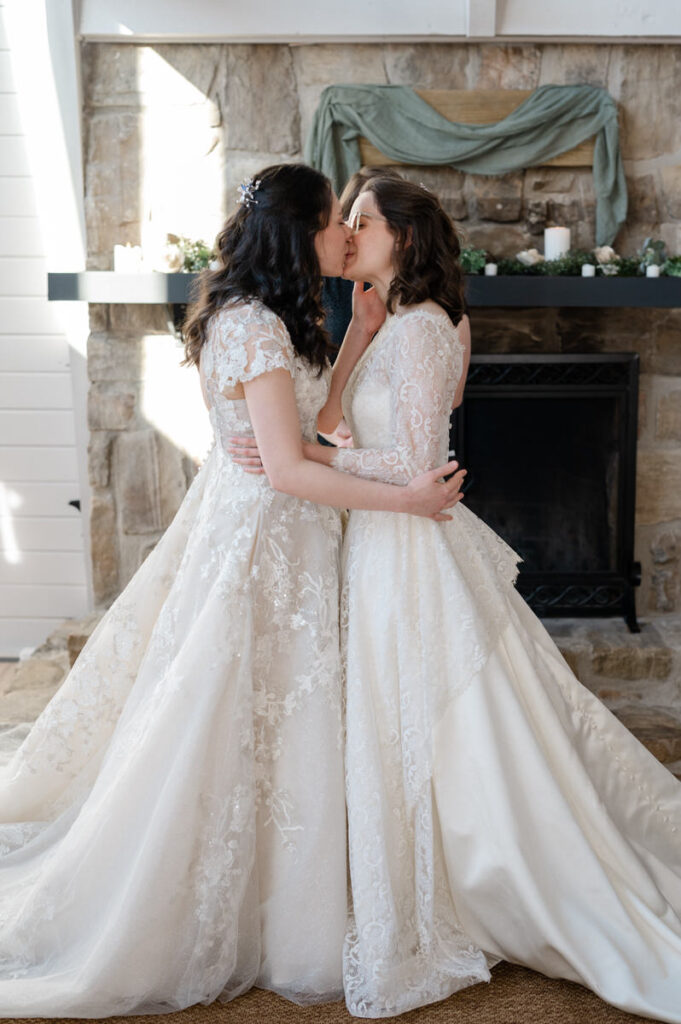 Two brides kissing in front of a fireplace. 
