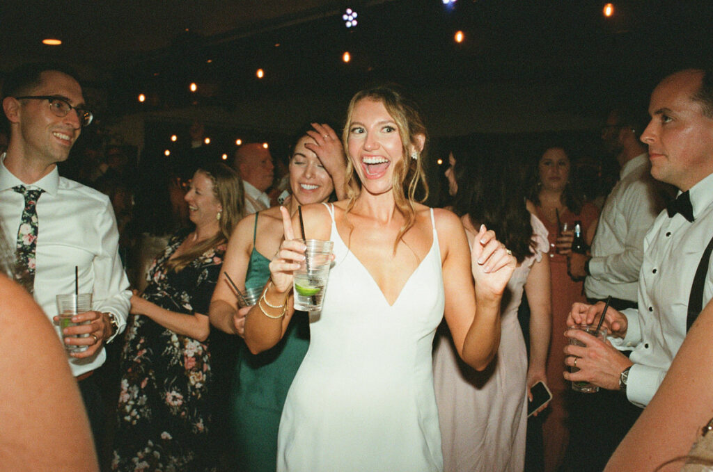 A happy bride holding a drink, surrounded by wedding guests, with a cheerful expression and a dance floor in the background.