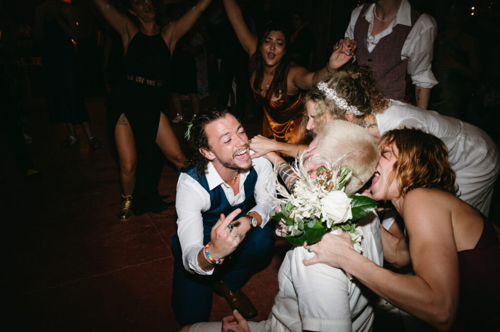 A joyous wedding reception scene with guests dancing and a man in a vest and tie kneeling and smiling at a bride holding a bouquet.
