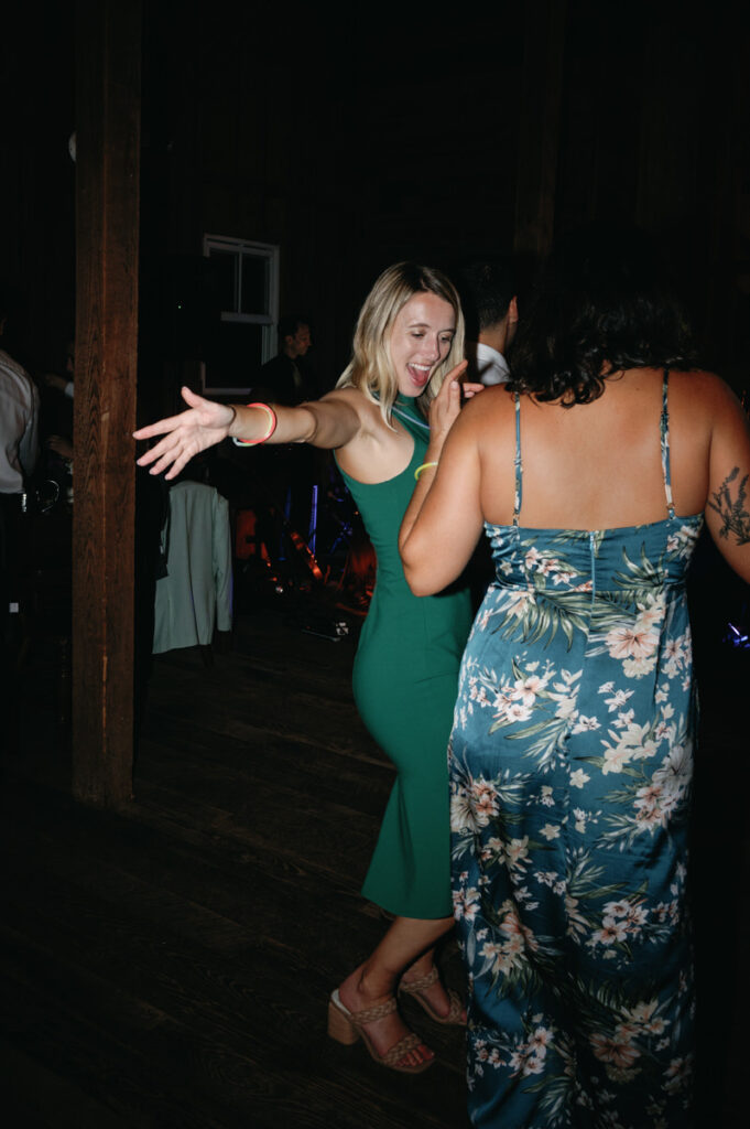 Two women dancing at a wedding reception, one in a green dress with arms open wide and the other in a floral dress facing away.