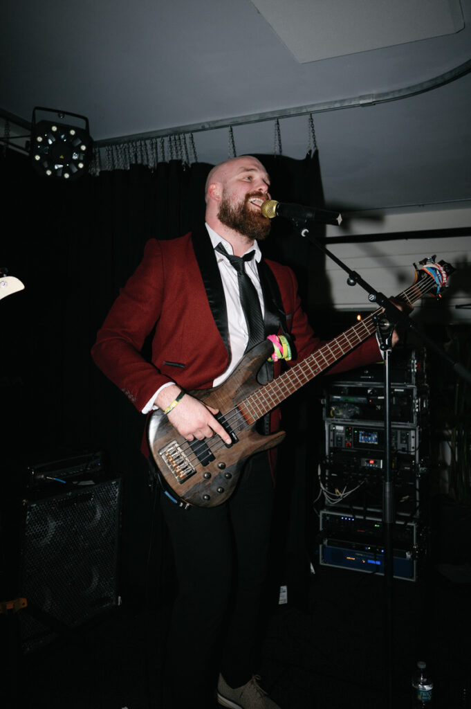 A bass guitarist in a red jacket and bow tie performing on stage at a wedding, singing into a microphone.