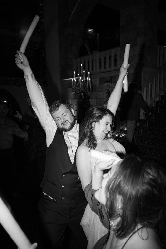 Black and white photo of wedding guests holding inflatable sticks in the air, dancing and celebrating with joy.