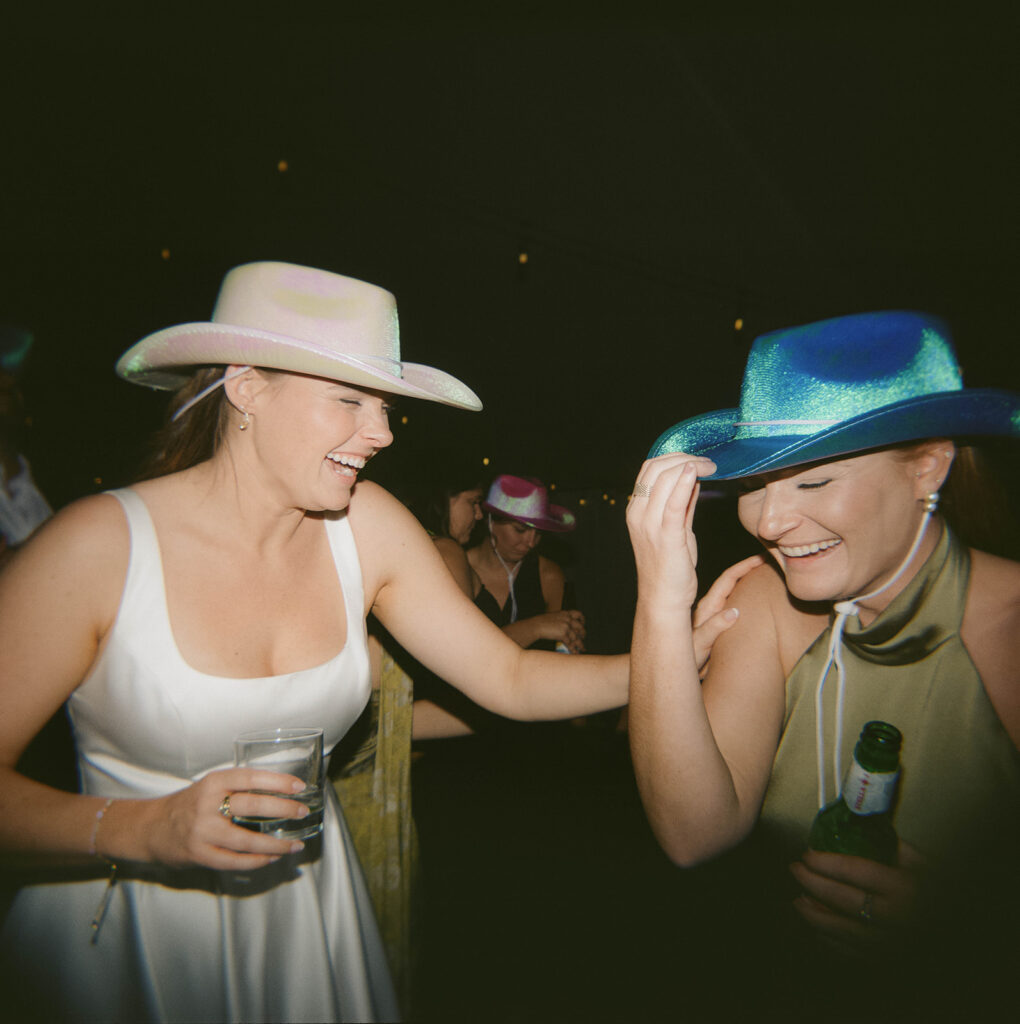 a bride and bridesmaid laugh together on the dance floor of a wedding. they both have drinks in hand and are wearing sparkly cowboy hats 