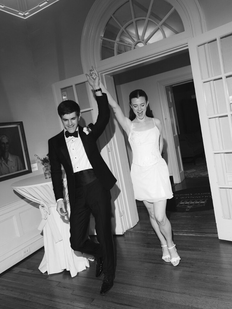 a black and white photo of a couple entering their reception with hands joined and raised overhead. the bride is wearing a mini dress while the groom is in a tuxedo 