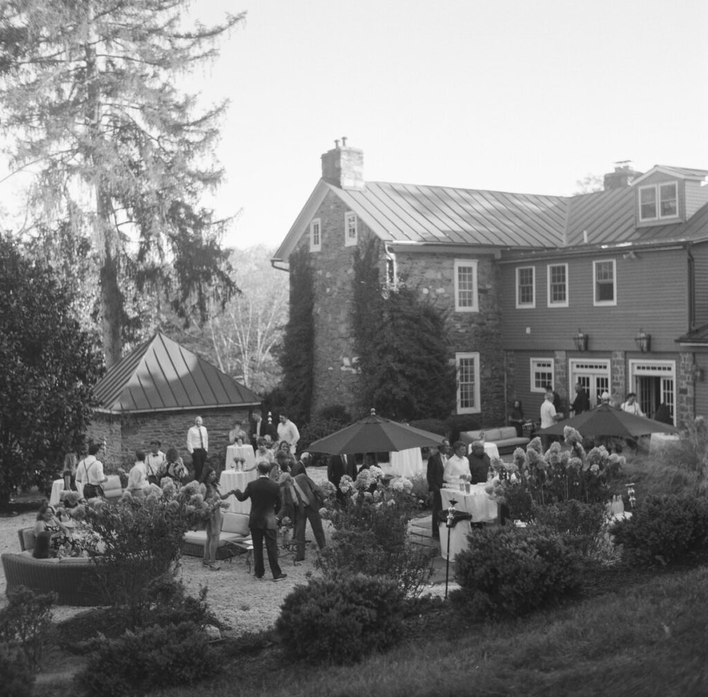A vintage black and white photo of guests mingling at an outdoor wedding reception with a historic stone house in the background