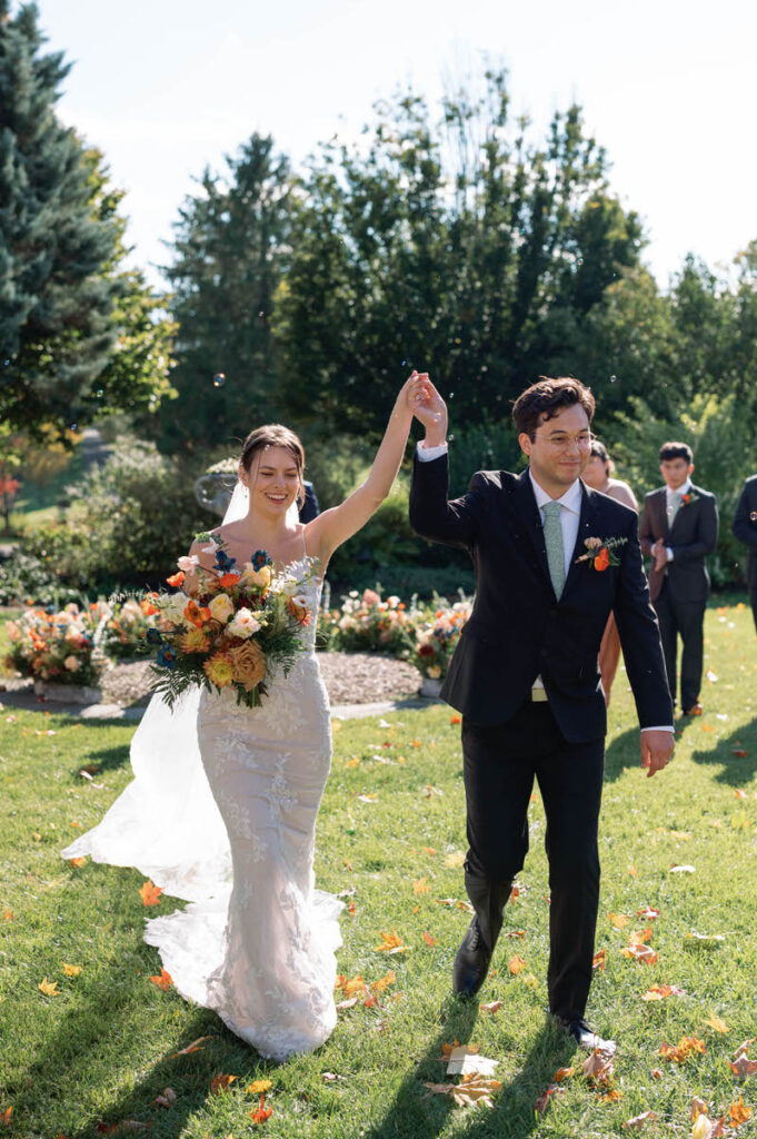 A newlywed couple holding hands in triumph as they walk down a garden aisle, with guests and autumn leaves surrounding them
