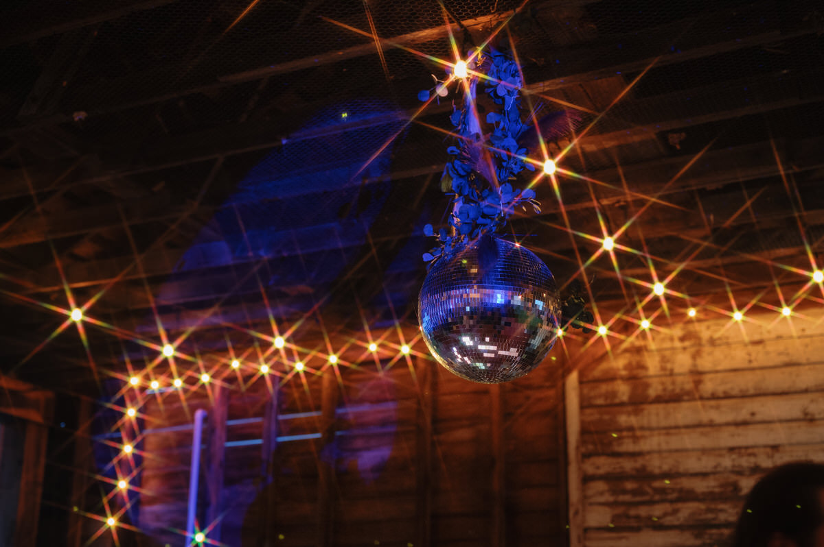 A disco ball adorned with blue flowers hanging from a rustic barn ceiling, casting sparkly light reflections around the room