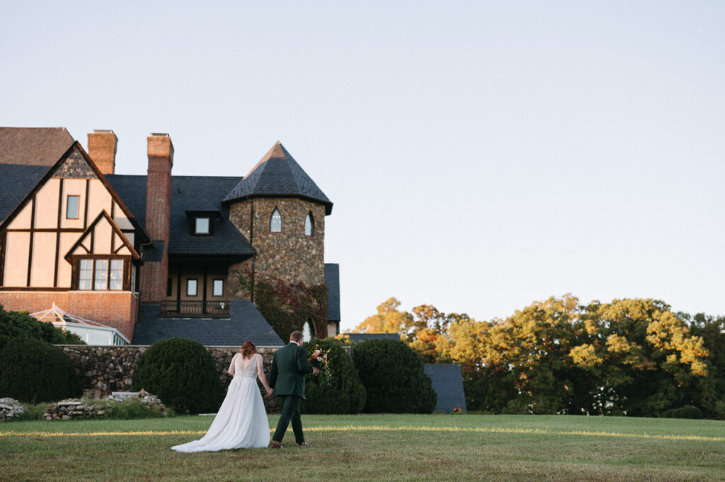 A couple in wedding attire walking away from the camera across a manicured lawn towards the grand Dover Hall, showcasing the elegant architecture of the building at dusk