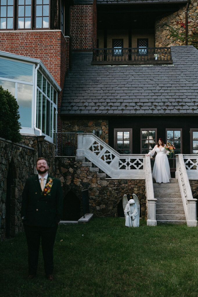 A groom in a green suit stands smiling on a lawn with a bride poised on a staircase behind him, set against the historical and ornate exterior of Dover Hall