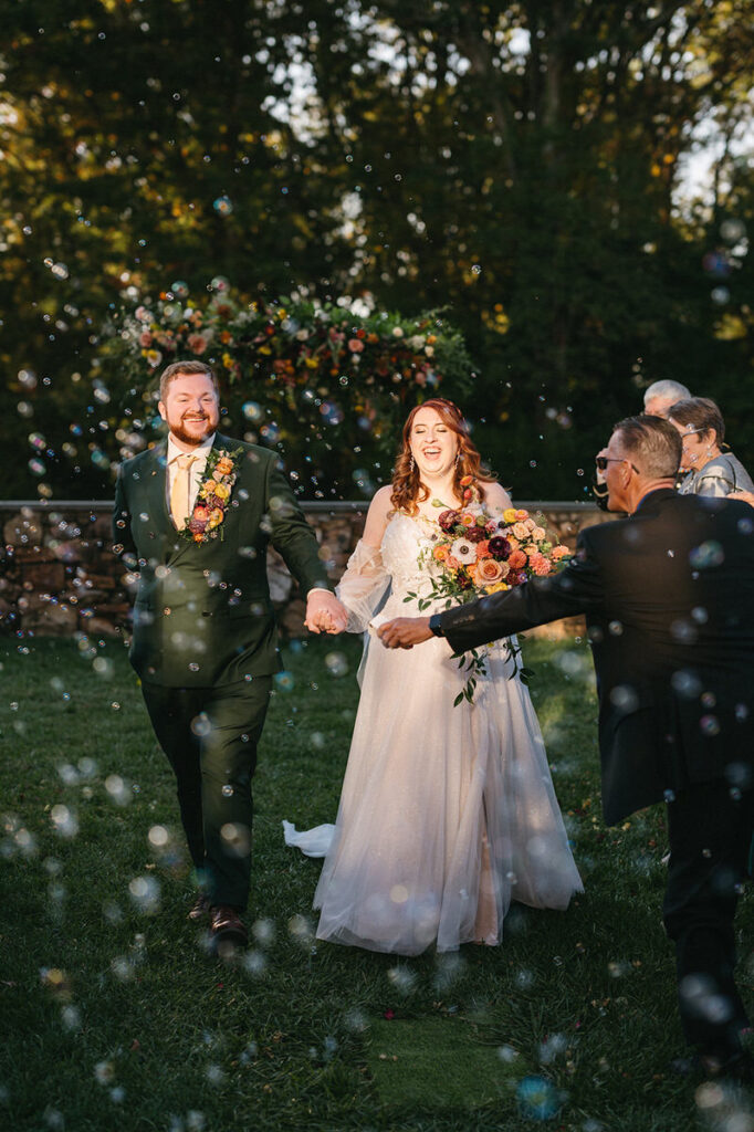 Bride and groom joyfully exit their wedding ceremony under a shower of bubbles, with guests and a floral arch in the background, capturing a moment of celebration.