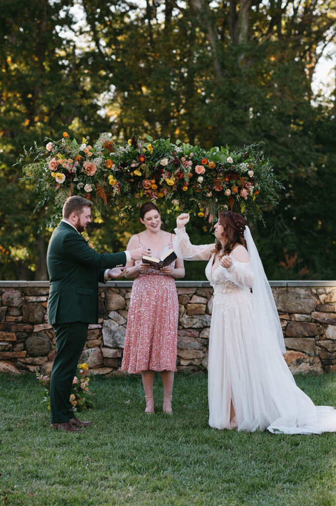 A wedding officiant reading from a book during a ceremony, with the bride and groom holding hands under a floral arch, sharing a light-hearted moment.