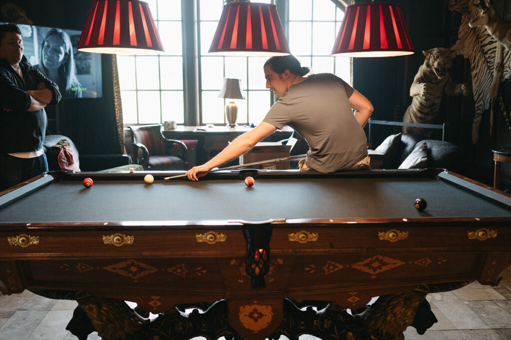 A man leans over an intricately carved pool table, focused on his shot, under the warm light of red lampshades, with a spectator standing arms crossed in a room adorned with taxidermy