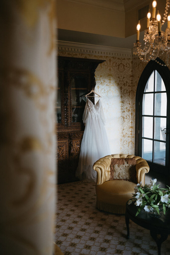 A bridal gown hanging in a window, with a vintage armchair and ornate decor in the room providing a glimpse into the bride's preparations at Dover Hall