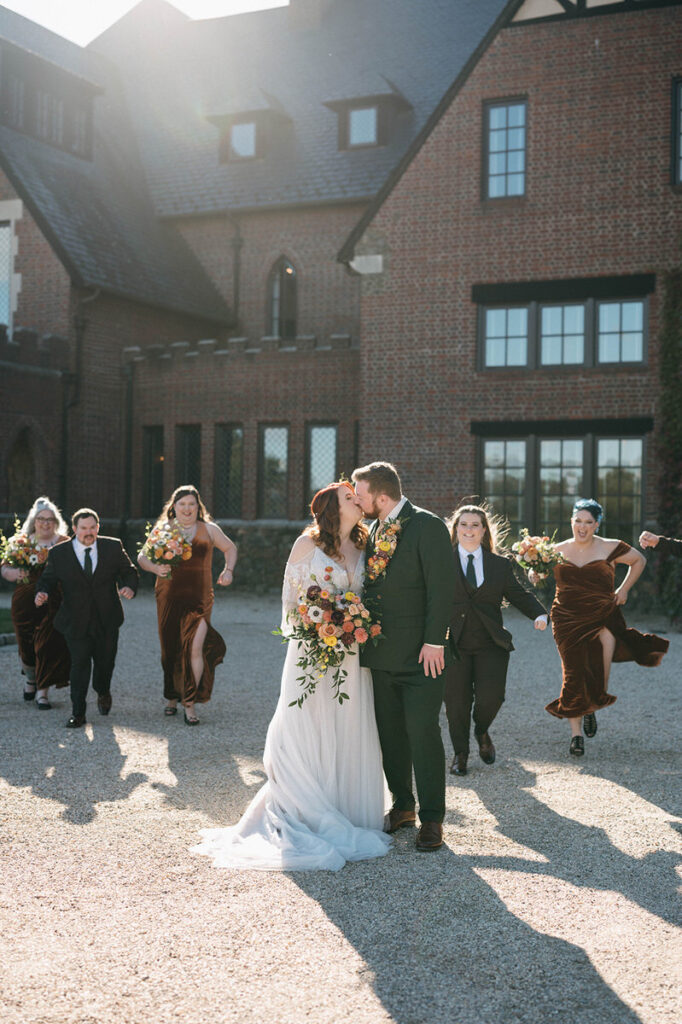 Bride and groom share a kiss on a gravel path with the bridal party in motion behind them, set against the stately brick facade of Dover Hall in the soft light of sunset