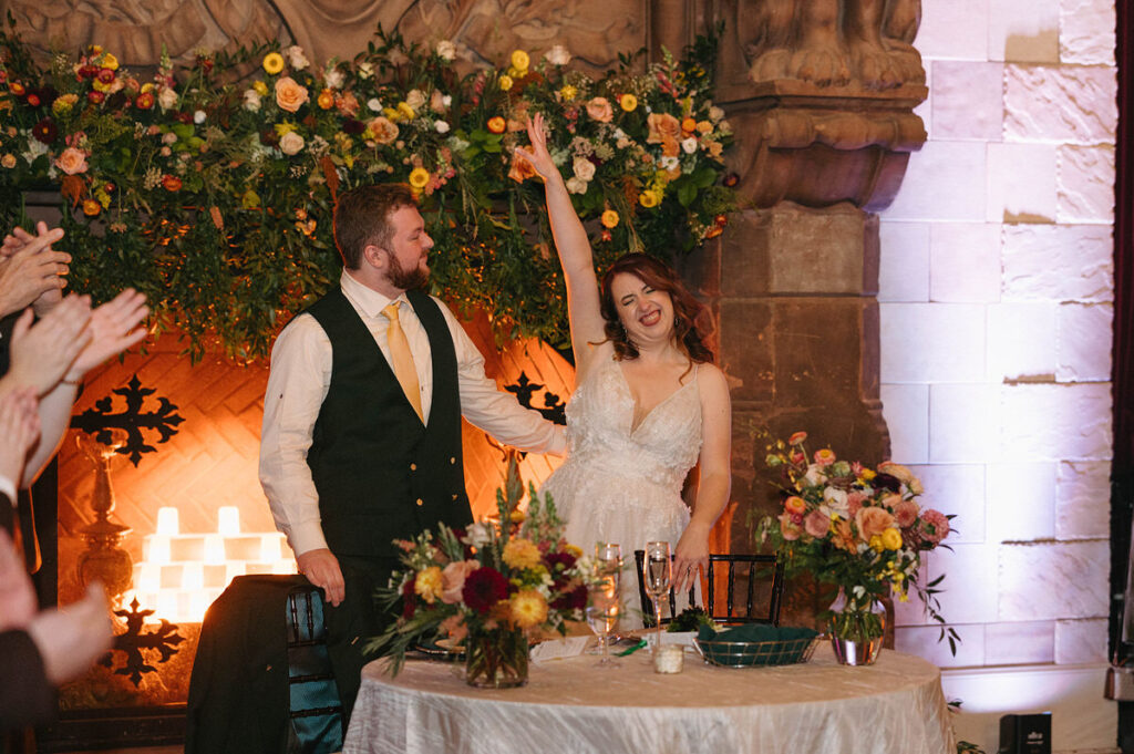 Bride and groom raise their hands in celebration at their wedding reception, surrounded by guests and a vibrant floral arrangement, with a romantic candlelit ambiance.