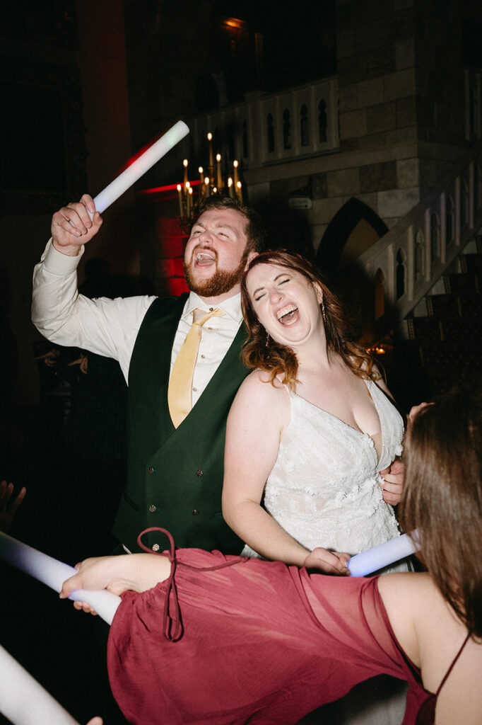 Bride and groom jubilantly celebrate, the groom wielding a light stick, surrounded by guests in a festive atmosphere with a hint of disco lighting