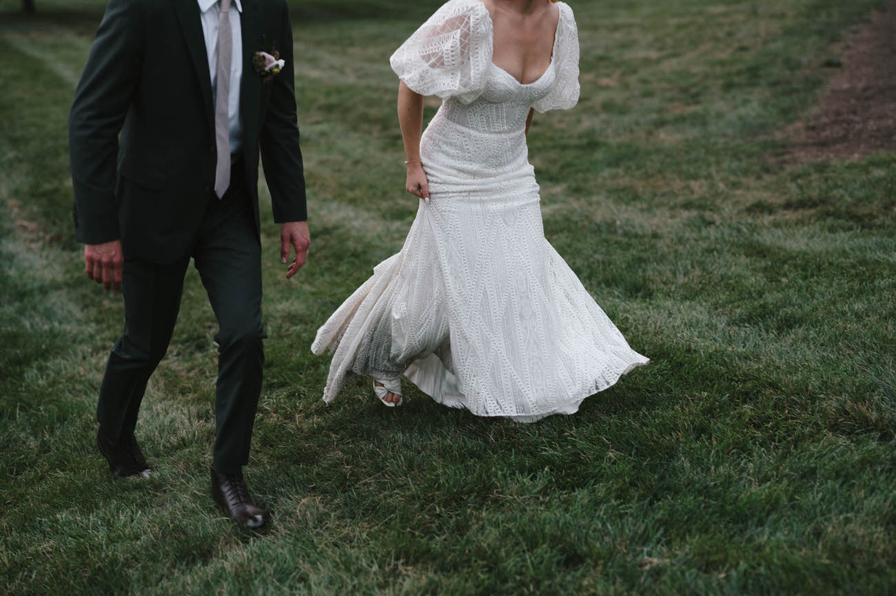 Bride in an elegant white lace gown and groom in a classic black suit walking side-by-side across a grassy field, with the focus on the couple's lower half and the bride's dress sweeping the ground.