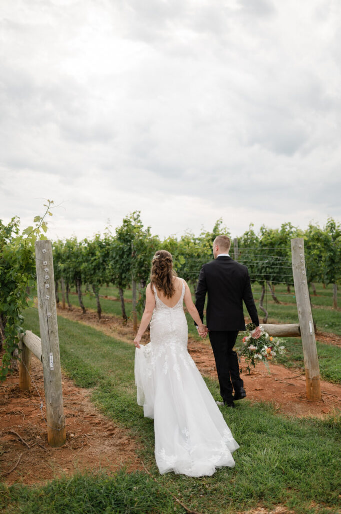 A bride and groom holding hands, seen from behind as they walk through a vineyard, with the bride carrying a bouquet and the trail of her white gown trailing on the ground.