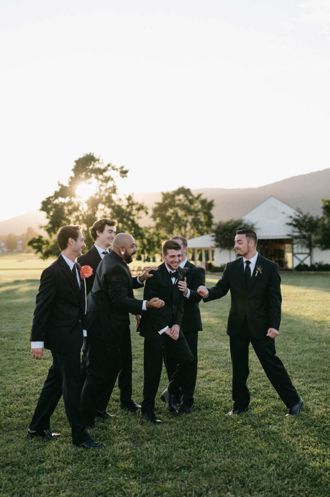 A group of groomsmen in black suits walking across a field, laughing and interacting with each other, with soft sunlight and mountains in the background.