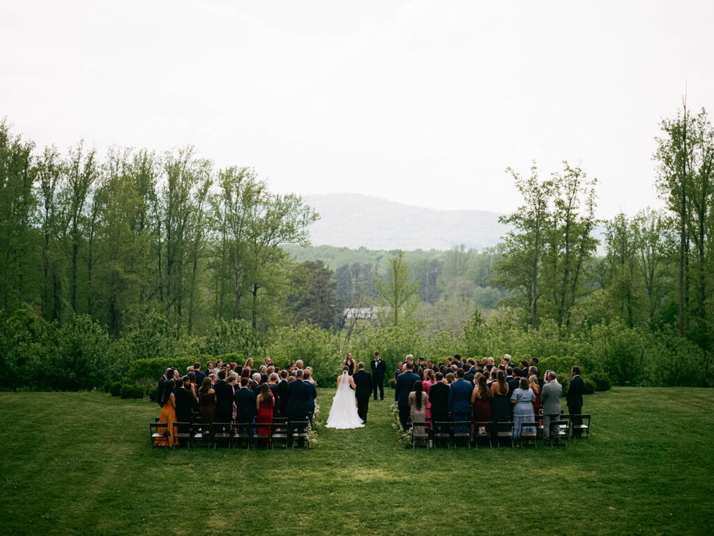 A bride and her father walk down the aisle to a wedding ceremony on a lush green lawn overlooking the blue ridge mountains at Eastwood Winery
