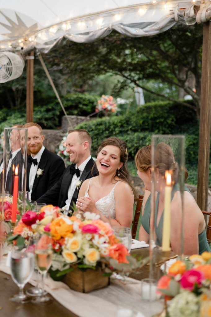 A bride laughing joyfully at the wedding reception table, surrounded by guests, with vibrant floral arrangements and lit candles in the foreground.