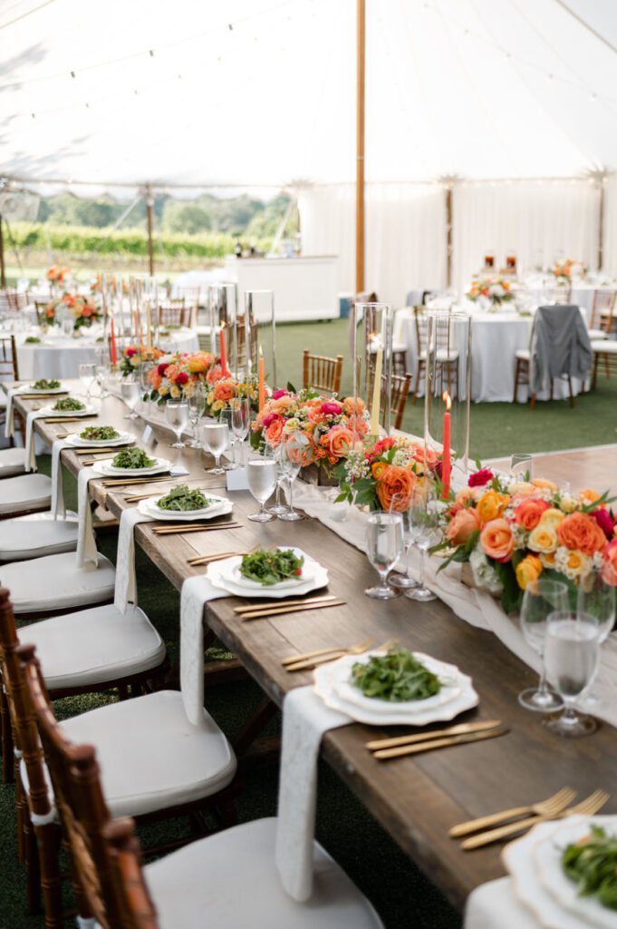 Elegantly arranged wedding reception tables with orange and pink floral centerpieces, set under a tent with a view of the vineyards in the distance.