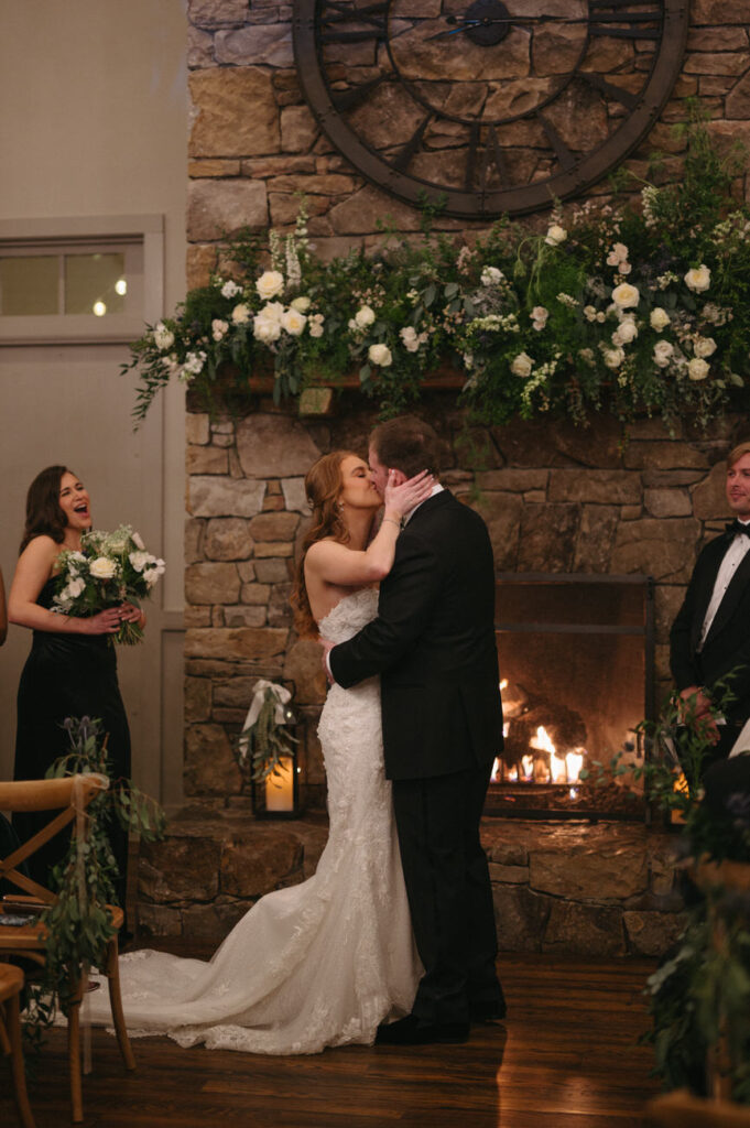 Bride and groom sharing a romantic kiss at the wedding altar with a stone fireplace and floral arrangements in the background, while guests look on.