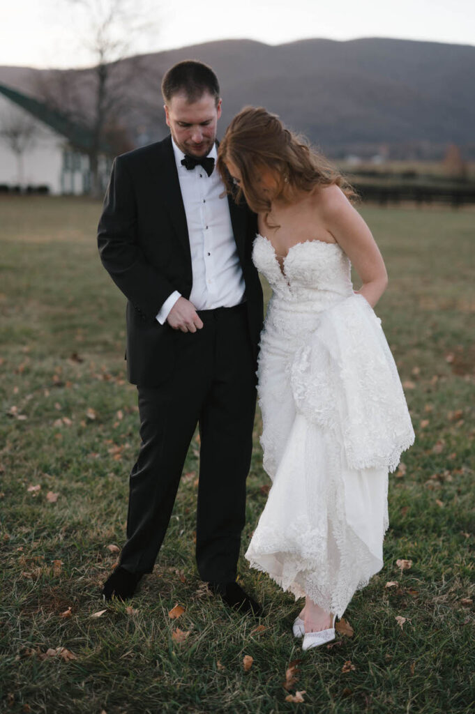 A contemplative moment between the bride and groom, with the bride holding her white lace gown while standing on a grassy field, and mountains in the backdrop.