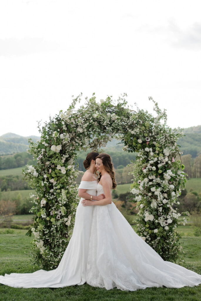 Two brides embracing in front of a lush floral archway, with the serene landscape of Virginia's countryside stretching out behind them.